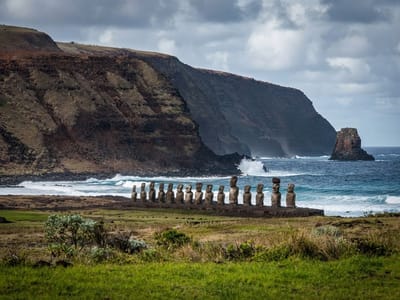 A row of stone Moai statues stands along a grassy shore against a backdrop of ocean waves and coastal cliffs.