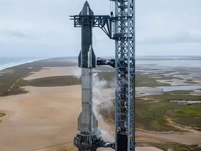 A tall two-stage rocket stands stacked on a launch mount next to a large metal launch tower with vapor venting along its side, set against a coastal landscape.