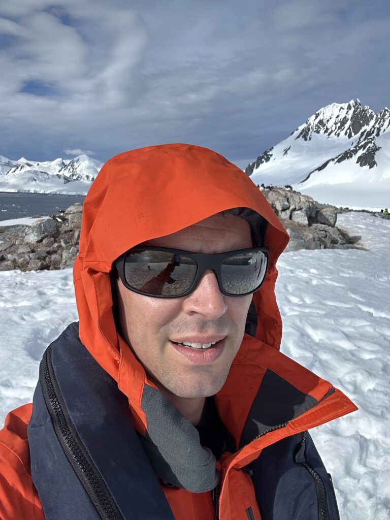 John Arrow taking a selfie in a red parka with snow-covered Antarctic mountains behind him