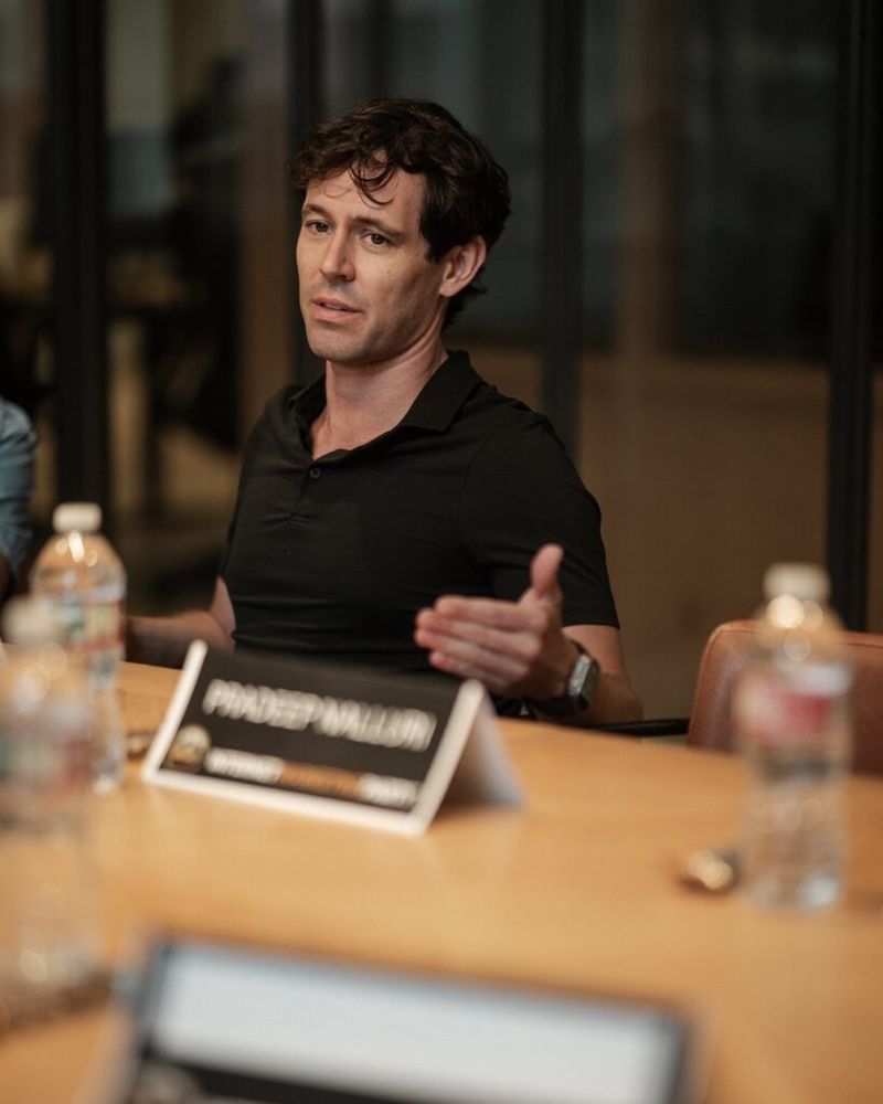 John Arrow speaking at a panel discussion, seated at a conference table with a name placard